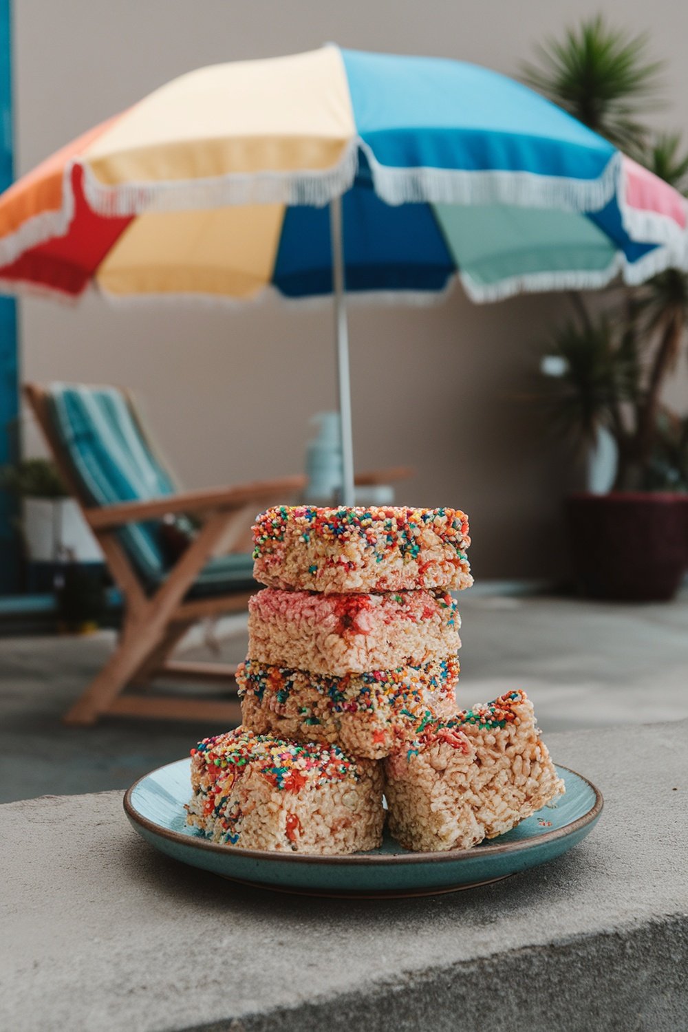 A stack of colorful crispy rice treats with sprinkles on a plate, with a beach umbrella in the background.